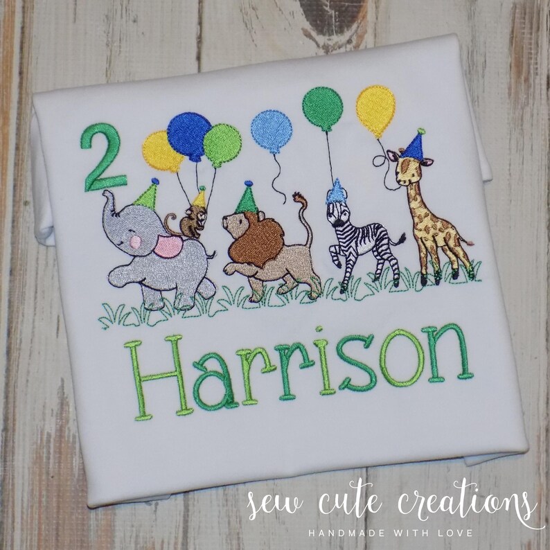 Puede incluir: Una camiseta blanca con un dise&ntilde;o bordado de un elefante gris, un le&oacute;n marr&oacute;n, una cebra blanca y negra y una jirafa, todos con gorros de fiesta. Los animales sostienen globos de colores. El n&uacute;mero 2 y el nombre Harrison est&aacute;n bordados debajo.