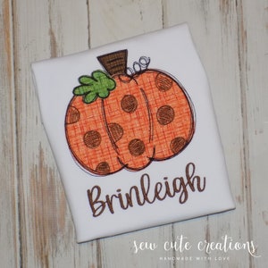 May include: A white t-shirt with a brown and orange pumpkin with brown polka dots and a green leaf. The name "Brinleigh" is embroidered in brown below the pumpkin. The text "sew cute creations" is embroidered in brown at the bottom of the shirt.