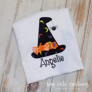 May include: White t-shirt with an embroidered witch hat, bow, and spider. The name "Angeli" is embroidered below the hat. The shirt has the text "sew cute creations handmade with love" at the bottom.