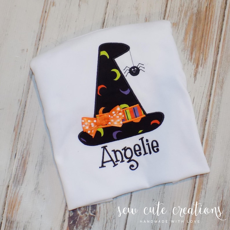May include: White t-shirt with an embroidered witch hat, bow, and spider. The name "Angeli" is embroidered below the hat. The shirt has the text "sew cute creations handmade with love" at the bottom.