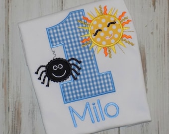 May include: White t-shirt with a blue gingham number one, a smiling sun, and a black spider. The name "Milo" is embroidered in blue. The sun is yellow with orange rays. The spider has a smiling face.