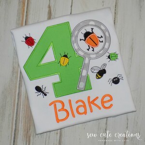 May include: White t-shirt with a green number 4 and the name "Blake" in orange. The shirt features embroidered insects, including a ladybug, bee, and spider, and a magnifying glass with a bug inside.