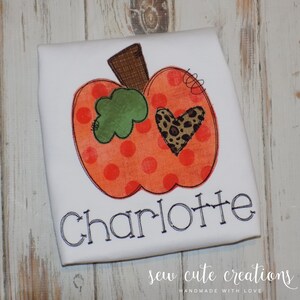 May include: White t-shirt with an orange pumpkin with red polka dots, a green leaf, and a brown stem. The pumpkin has a brown and black leopard print heart on it. The name "Charlotte" is embroidered below the pumpkin. The text "sew cute creations" is at the bottom of the shirt.