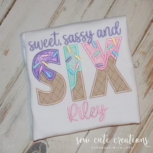 May include: White t-shirt with the text "sweet, sassy and SIX" in colorful embroidered letters that look like ice cream cones. The name "Riley" is embroidered below the number in pink thread.