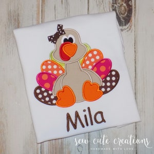 Puede incluir: Camiseta blanca con un pavo marrón bordado con plumas de lunares rosas, naranjas y blancos. El pavo tiene un lazo marrón y el nombre "Mila" está bordado debajo del pavo.