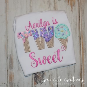 May include: A white t-shirt with pink, purple and blue embroidered ice cream cones that spell out "TWO" and the word "Sweet" in pink embroidery. The text "Aerilyn is" is embroidered above the ice cream cones.