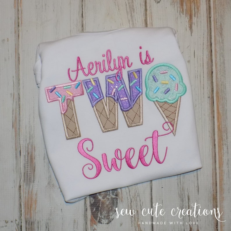 May include: A white t-shirt with pink, purple and blue embroidered ice cream cones that spell out "TWO" and the word "Sweet" in pink embroidery. The text "Aerilyn is" is embroidered above the ice cream cones.