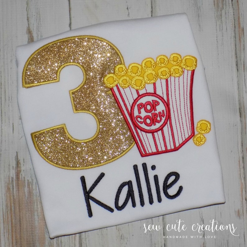 May include: A white t-shirt with a gold glitter number 3 and a red and white popcorn bucket with yellow popcorn. The name "Kallie" is written below the number and popcorn bucket. The text "sew cute creations handmade with love" is at the bottom of the shirt.
