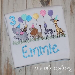 Puede incluir: Una camiseta blanca con un dise&ntilde;o bordado de un elefante gris, un mono, un le&oacute;n, una cebra y una jirafa, todos con gorros de fiesta y sosteniendo globos. El n&uacute;mero 3 y el nombre Emmie est&aacute;n bordados con hilo de color azul.