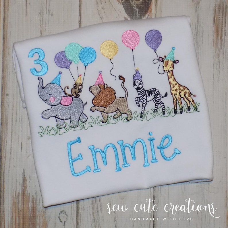 Puede incluir: Una camiseta blanca con un dise&ntilde;o bordado de un elefante gris, un mono, un le&oacute;n, una cebra y una jirafa, todos con gorros de fiesta y sosteniendo globos. El n&uacute;mero 3 y el nombre Emmie est&aacute;n bordados con hilo de color azul.