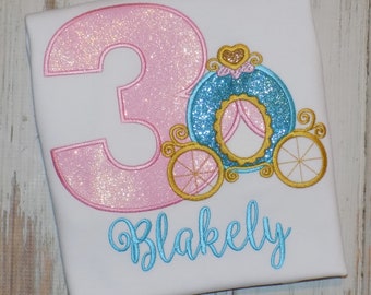 May include: A white t-shirt featuring an embroidered design of a large pink glittery number three next to a blue glittery pumpkin carriage with gold wheels. The name Blakely is embroidered below in light blue cursive script lettering.