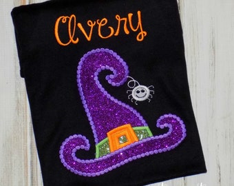 May include: Black t-shirt with an embroidered purple glitter witch hat, orange buckle, and a smiling spider. The name "Avery" is embroidered in orange above the hat. The words "sew cute creations" are at the bottom.