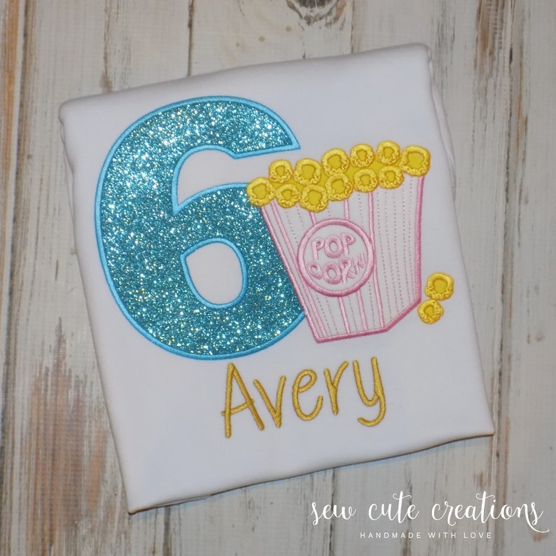 May include: A white t-shirt with a blue glitter number 6, a pink and white popcorn bucket with yellow popcorn, and the name Avery in yellow thread. The shirt says "Sew Cute Creations Handmade with Love".