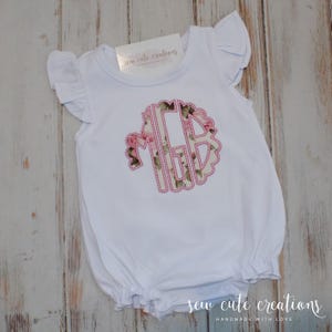May include: White baby romper with ruffled sleeves and hem. Features a floral monogram in pink and green, stitched onto the front. The romper has the text "sew cute creations" and "handmade with love".