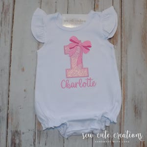 May include: White baby romper with pink glittery number 1 and bow design. The name "Charlotte" is embroidered below the number. The romper has ruffled sleeves and the text "sew cute creations" is visible.