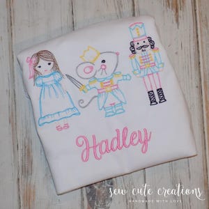 May include: White t-shirt with embroidered images of a girl, a mouse, and a nutcracker. The name "Hadley" is embroidered in pink below the images. The text "sew cute creations handmade with love" is printed at the bottom of the shirt.