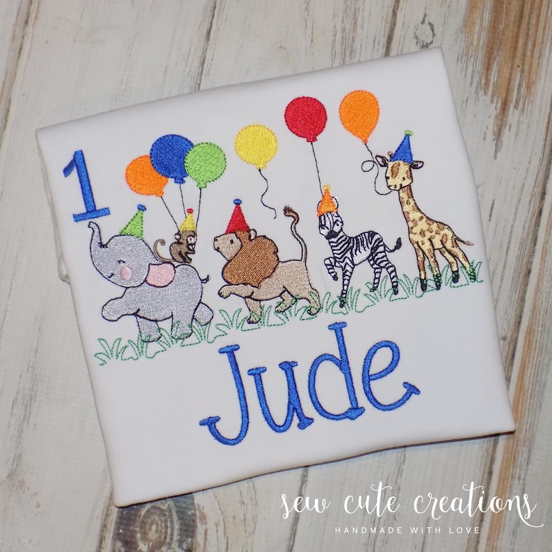 Puede incluir: Una camiseta blanca con un dise&ntilde;o bordado de un elefante gris, un le&oacute;n marr&oacute;n, una cebra blanca y negra y una jirafa, todos con gorros de fiesta coloridos. Sostienen globos y el n&uacute;mero 1, con el nombre Jude bordado en color azul.