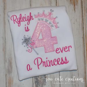 May include: White t-shirt with pink embroidered text and design. The text reads "Ryleigh is 4 ever a Princess." A pink glittery number 4 is centered, topped with a crown. A silver wand is to the left of the number.