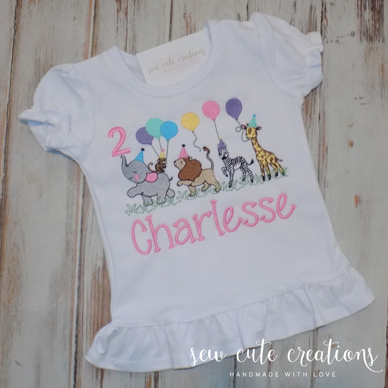 Puede incluir: Una camiseta blanca de manga corta con dobladillo de volantes que presenta un dise&ntilde;o bordado de un elefante gris, un mono, un le&oacute;n, una cebra y una jirafa con gorros de fiesta. El n&uacute;mero 2 y el nombre Charlesses est&aacute;n bordados en rosa.
