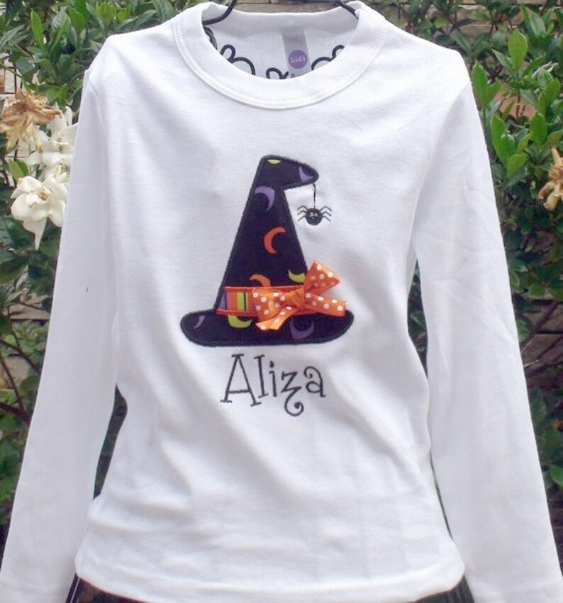 May include: White long-sleeved shirt with an embroidered witch hat, orange and white polka dot bow, and spider. The shirt is personalized with the name "Aliza".