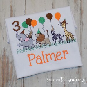 Puede incluir: Una camiseta blanca con un dise&ntilde;o bordado de un elefante gris, un le&oacute;n marr&oacute;n, una cebra y una jirafa, todos con gorros de fiesta. El dise&ntilde;o incluye el n&uacute;mero 3, globos de colores, hierba verde y el nombre Palmer en texto de color naranja.