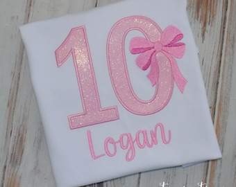 May include: A white t-shirt featuring a large embroidered number 10 in pink glitter fabric. The number zero is accented with a pink fabric bow. Below the number, the name Logan is embroidered in pink cursive script lettering.