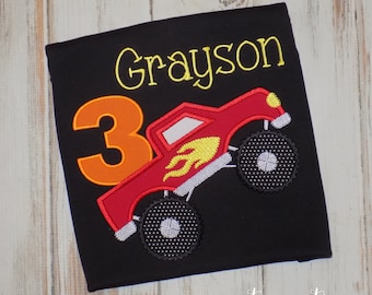 May include: Black t-shirt featuring a red monster truck applique with yellow flames and the number 3 in orange. The name "Grayson" is embroidered in yellow. The text "sew cute creations" is at the bottom.
