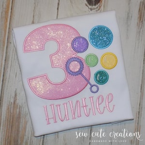 May include: White t-shirt with a pink glittery number 3 and colorful bubble design. The name "Huntlee" is embroidered in pink script below the number. The shirt is decorated with a bubble wand.