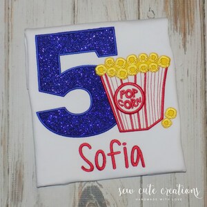 May include: A white t-shirt with a blue glittery number 5 and a red and white popcorn bucket with yellow popcorn. The name "Sofia" is embroidered in red below the number and popcorn. The text "sew cute creations" is embroidered in black at the bottom of the shirt.