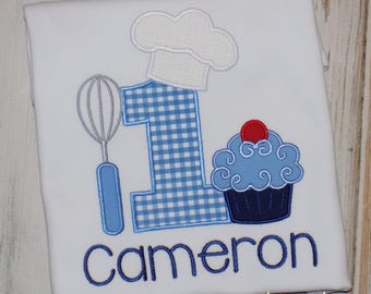 May include: A white t-shirt featuring an embroidered design of a blue gingham number one topped with a white chef hat. To the left is a whisk, and to the right is a blue cupcake with a red cherry. The name Cameron is embroidered in blue below.