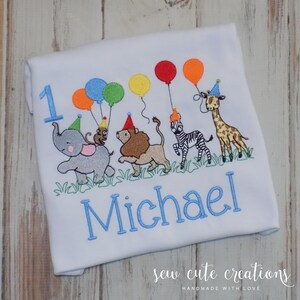 Puede incluir: Una camiseta blanca con un dise&ntilde;o bordado de un elefante gris, un le&oacute;n marr&oacute;n, una cebra y una jirafa con gorros de fiesta. Los animales sostienen globos de colores. El n&uacute;mero 1 y el nombre Michael est&aacute;n bordados en color azul.