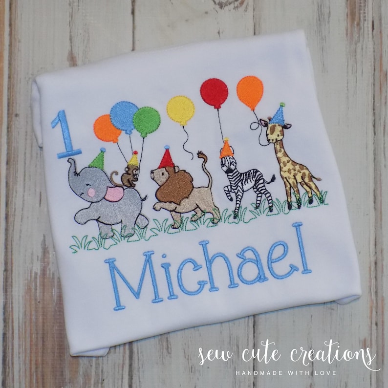Puede incluir: Una camiseta blanca con un dise&ntilde;o bordado de un elefante gris, un le&oacute;n marr&oacute;n, una cebra y una jirafa con gorros de fiesta. Los animales sostienen globos de colores. El n&uacute;mero 1 y el nombre Michael est&aacute;n bordados en color azul.