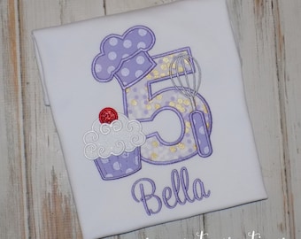 May include: A white t-shirt featuring an embroidered design of the number five in purple with gold polka dots, topped with a purple chef hat. A cupcake with a red cherry and a silver whisk are attached to the number. The name Bella is embroidered below.