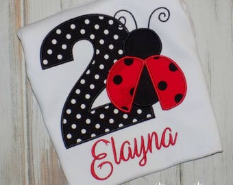 May include: A white t-shirt with a black and white polka dot number two and a red ladybug with black spots. The name "Elayna" is embroidered in red below the number. The shirt is handmade with love and is perfect for a child's second birthday.