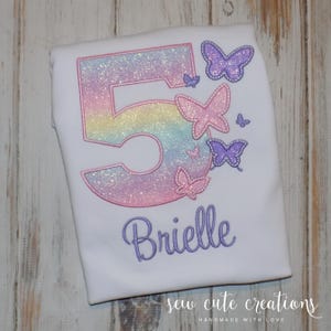 Puede incluir: Una camiseta blanca con un número 5 de purpurina arcoíris y mariposas bordadas. El nombre "Brielle" está bordado debajo del número. La camiseta es para el cumpleaños de un niño.