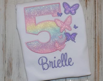 May include: A white t-shirt with a rainbow glitter number 5 and butterflies embroidered on it. The name "Brielle" is embroidered below the number. The shirt is for a child's birthday.