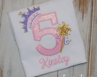 Puede incluir: Número cinco de purpurina rosa con una corona morada y una varita de purpurina dorada. El nombre Kinsley está bordado debajo del número. El diseño está en una camiseta blanca.
