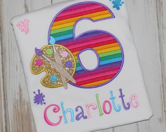 May include: A white t-shirt with a rainbow striped number six, a gold glitter paint palette with paintbrushes, and the name Charlotte embroidered in colorful letters. The shirt is decorated with paint splatters.