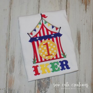 Puede incluir: Un pelele blanco con un diseño de carpa de circo roja, blanca y azul. La carpa tiene un círculo amarillo con un número uno blanco en el centro. El nombre "IKER" está bordado debajo de la carpa en letras rojas, amarillas, verdes y azules. El texto "sew cute creations handmade with love" está en la parte inferior del pelele.