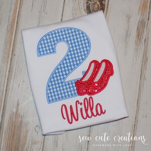May include: A white t-shirt with a blue and white gingham number 2, red glitter ruby slippers, and the name "Willa" embroidered in red. The shirt is for a child's birthday.