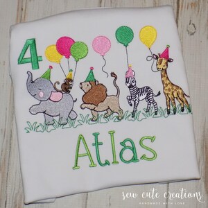 Puede incluir: Una camiseta blanca con un dise&ntilde;o bordado que incluye el n&uacute;mero 4 en verde, el nombre Atlas en verde y un grupo de animales de safari como un elefante, mono, le&oacute;n, cebra y jirafa con gorros de fiesta y globos de colores.
