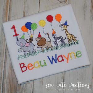 Puede incluir: Una camiseta blanca con un dise&ntilde;o bordado de un elefante gris, un le&oacute;n marr&oacute;n, una cebra blanca y negra y una jirafa, todos con gorros de fiesta. Los animales sostienen globos de colores. El n&uacute;mero 1 y el nombre Beau Wayne est&aacute;n bordados con hilo multicolor.