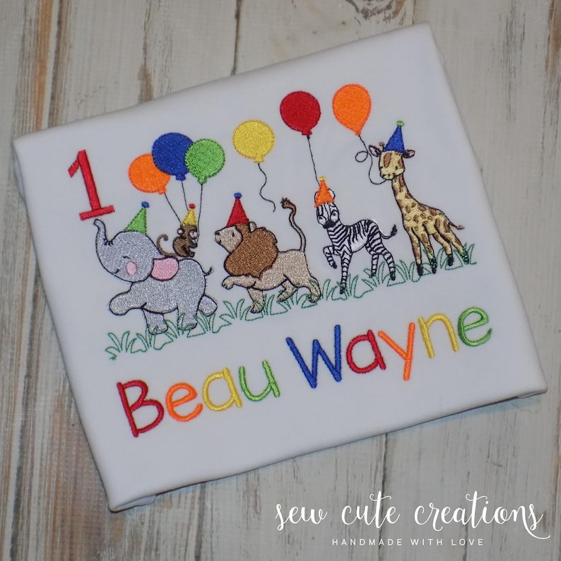 Puede incluir: Una camiseta blanca con un dise&ntilde;o bordado de un elefante gris, un le&oacute;n marr&oacute;n, una cebra blanca y negra y una jirafa, todos con gorros de fiesta. Los animales sostienen globos de colores. El n&uacute;mero 1 y el nombre Beau Wayne est&aacute;n bordados con hilo multicolor.