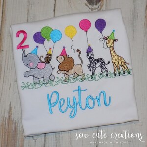 Puede incluir: Una camiseta blanca con un dise&ntilde;o bordado de un elefante gris, un le&oacute;n marr&oacute;n, una cebra blanca y negra y una jirafa, todos con gorros de fiesta coloridos. El dise&ntilde;o incluye el n&uacute;mero 2, globos y el nombre Peyton en color azul.