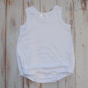 Puede incluir: Un pelele blanco liso sin mangas con cuello redondo, dos botones decorativos en los tirantes y cierre de corchetes en la parte inferior. La prenda cuenta con aberturas el&aacute;sticas en las piernas y un dise&ntilde;o sencillo y limpio.