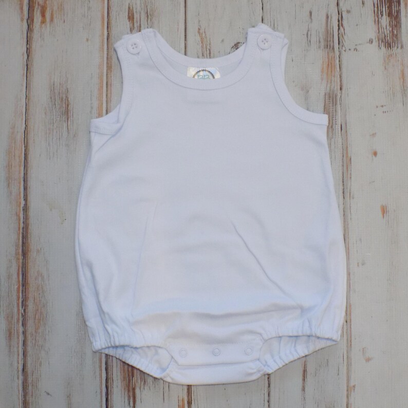 Puede incluir: Un pelele blanco liso sin mangas con cuello redondo, dos botones decorativos en los tirantes y cierre de corchetes en la parte inferior. La prenda cuenta con aberturas el&aacute;sticas en las piernas y un dise&ntilde;o sencillo y limpio.