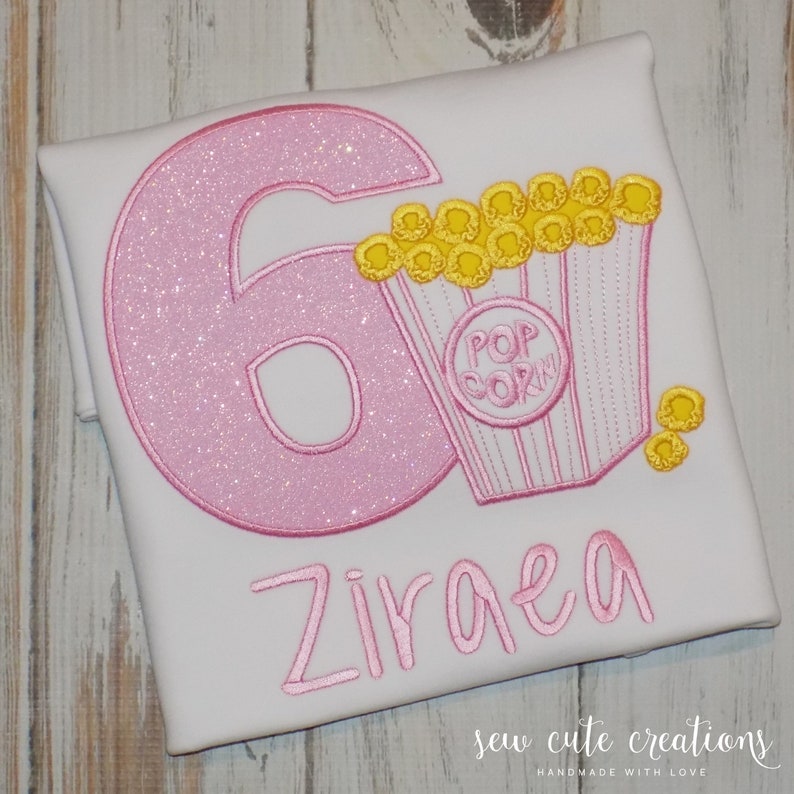 May include: Pink glitter number six with a pink and yellow popcorn bucket and the name Ziraea embroidered on a white t-shirt.