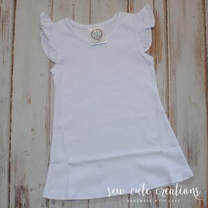 Puede incluir: Un vestido blanco liso de manga corta con volantes en los hombros y cuello redondo. Una peque&ntilde;a etiqueta circular de marca es visible en la parte interior del cuello, mostrando el texto BB y la talla 12M en una fuente clara y legible.
