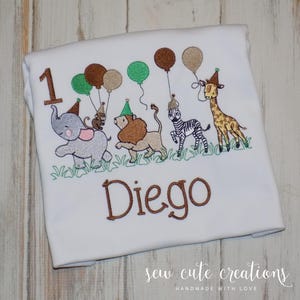 Puede incluir: Una camiseta blanca con un dise&ntilde;o bordado del n&uacute;mero 1 en marr&oacute;n, un elefante gris, un mono marr&oacute;n, un le&oacute;n beige, una cebra blanca y negra y una jirafa amarilla, todos con gorros de fiesta y globos, con el nombre Diego bordado debajo.