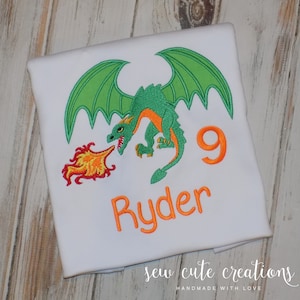 May include: A white t-shirt featuring an embroidered green dragon with orange accents breathing fire. The design includes the orange embroidered number 9 and the name Ryder in orange lettering. The embroidery is centered on the front of the shirt.